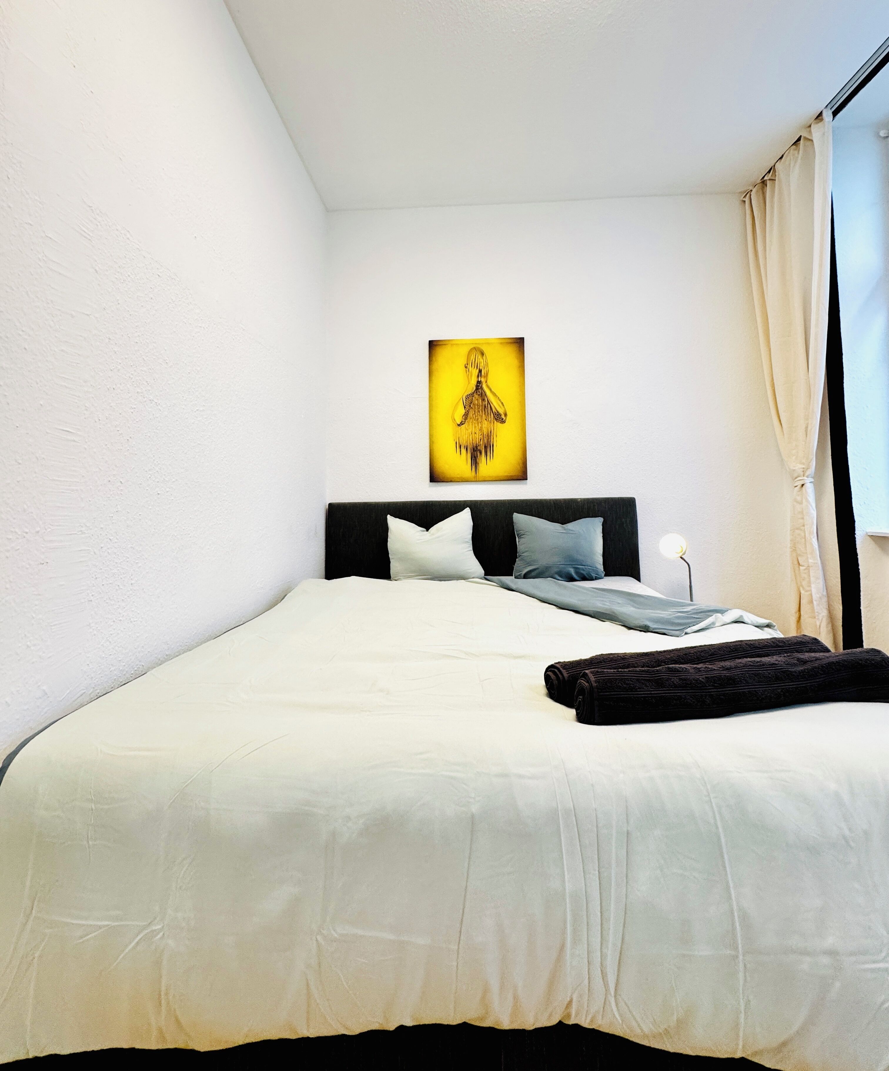 Ein ordentlich bezogenes Bett mit weißer Daunendecke, dunkelbraunem Kopfteil und einem einzelnen Kissen. Ein gerahmtes Kunstwerk hängt an der Wand darüber.