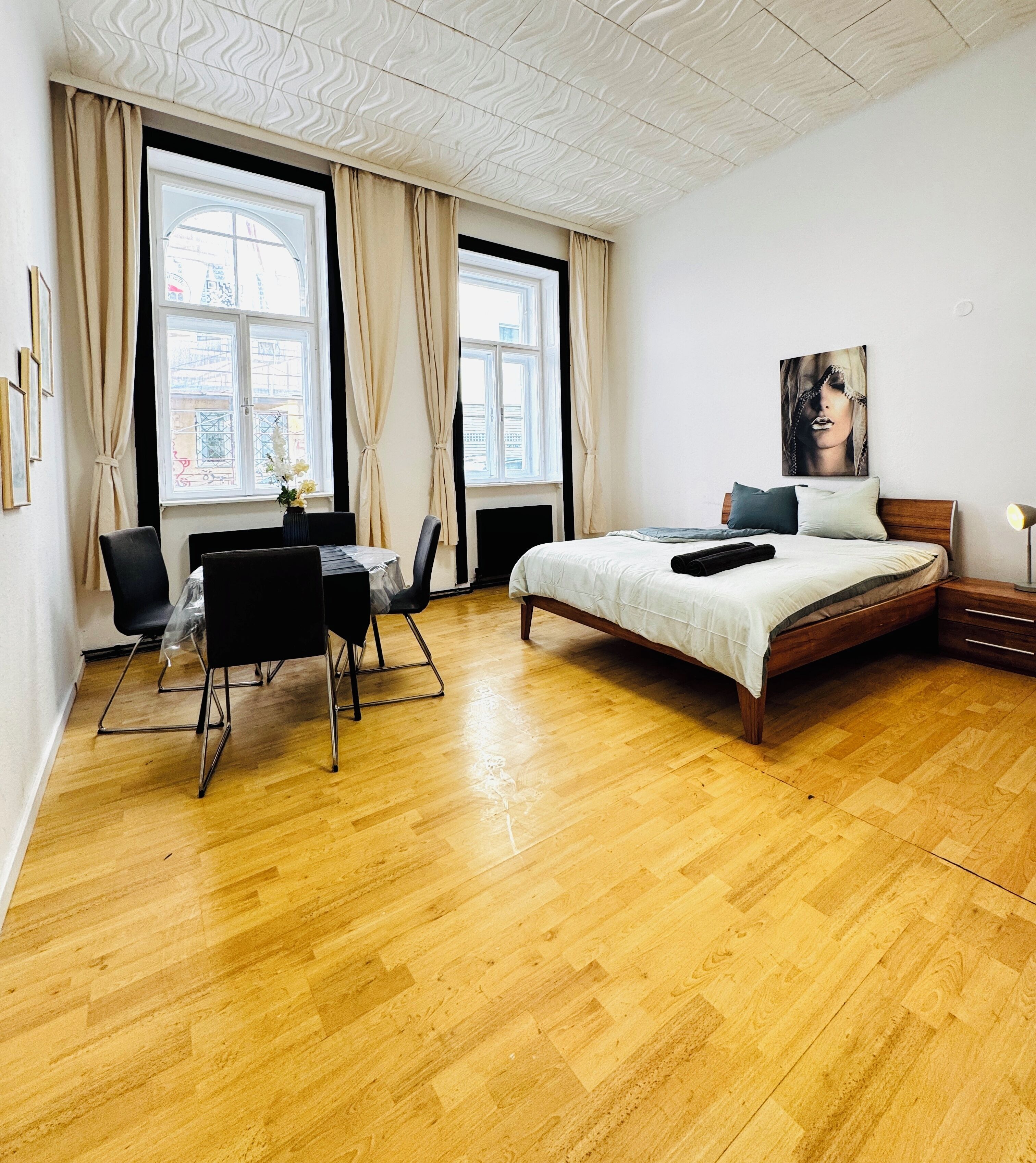 A bedroom with a wooden floor, a bed with a wooden frame, a desk with a chair, and large windows with curtains.