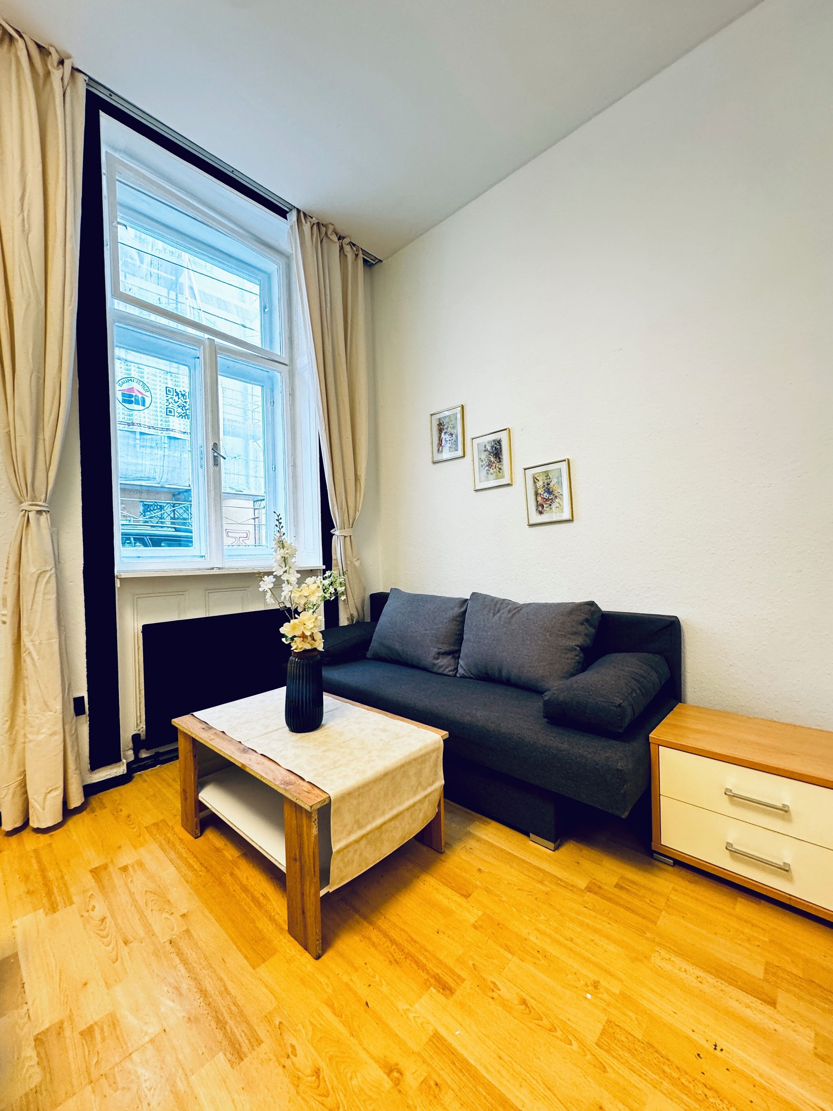 Ein Wohnzimmer mit einer Couch, einem kleinen Tisch mit einer Vase, einem Heizkörper und einem Fenster mit Vorhängen.