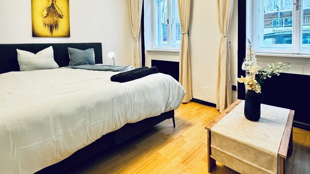 Ein Hotelzimmer mit einem großen Bett, einem Nachttisch mit Blumen in einer Vase, einer Deckenleuchte und einem Fenster mit Vorhängen.