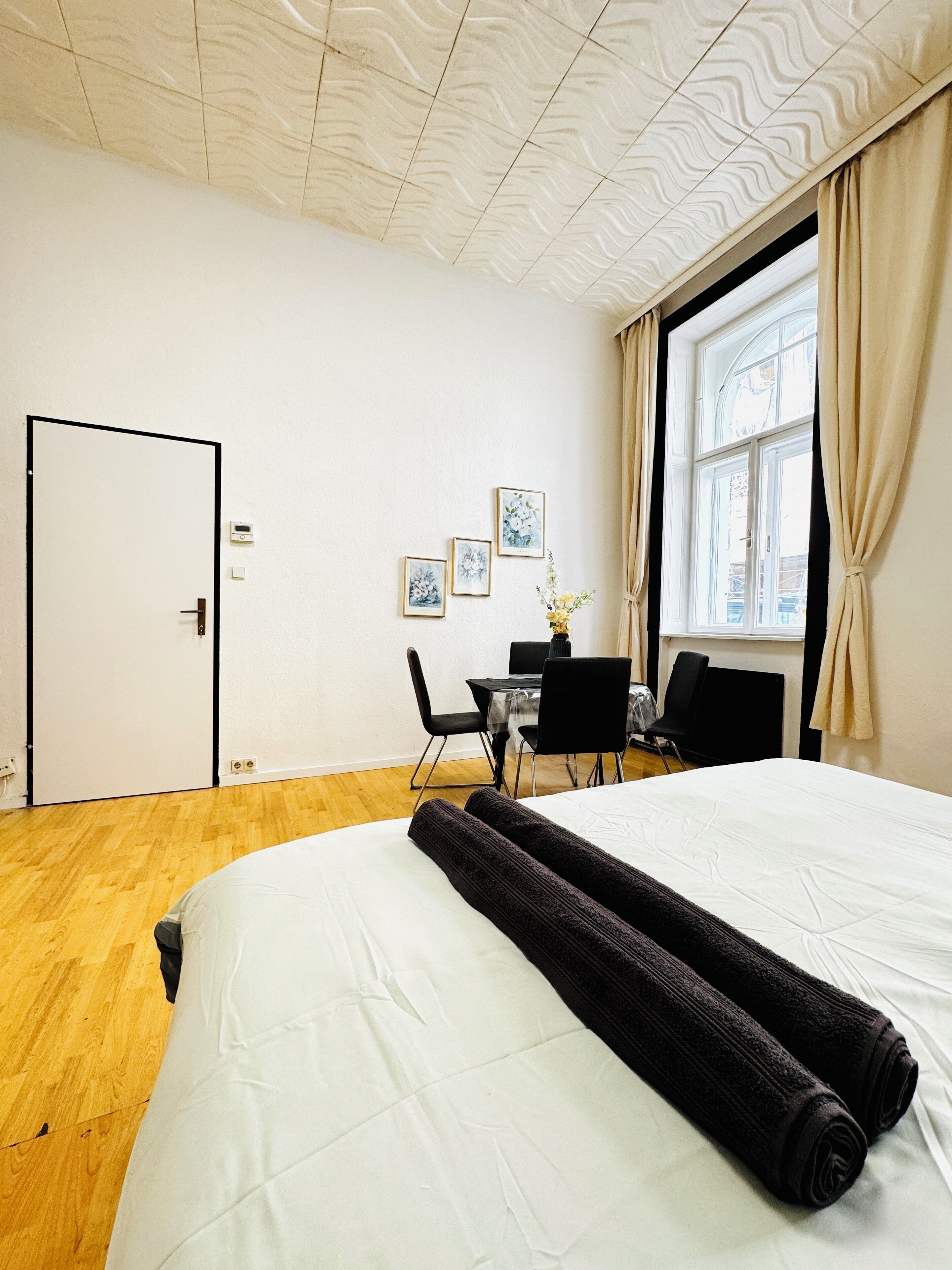 A hotel room with a bed, a desk, two chairs, a window with curtains, and three framed pictures on the wall.