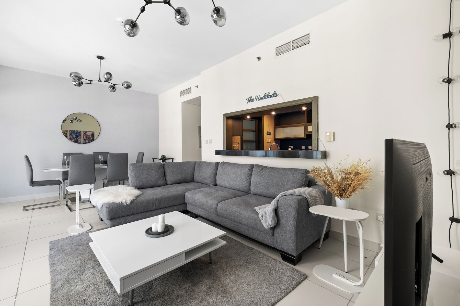 Een moderne woonkamer met een grijze modulaire bank, een witte salontafel, een eethoek met een ronde spiegel, en een keuken met een bordje waarop 'The Hardwoods' staat.