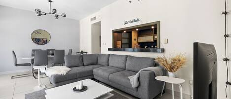 Een moderne woonkamer met een grijze modulaire bank, een witte salontafel, een eethoek met een ronde spiegel, en een keuken met een bordje waarop 'The Hardwoods' staat.