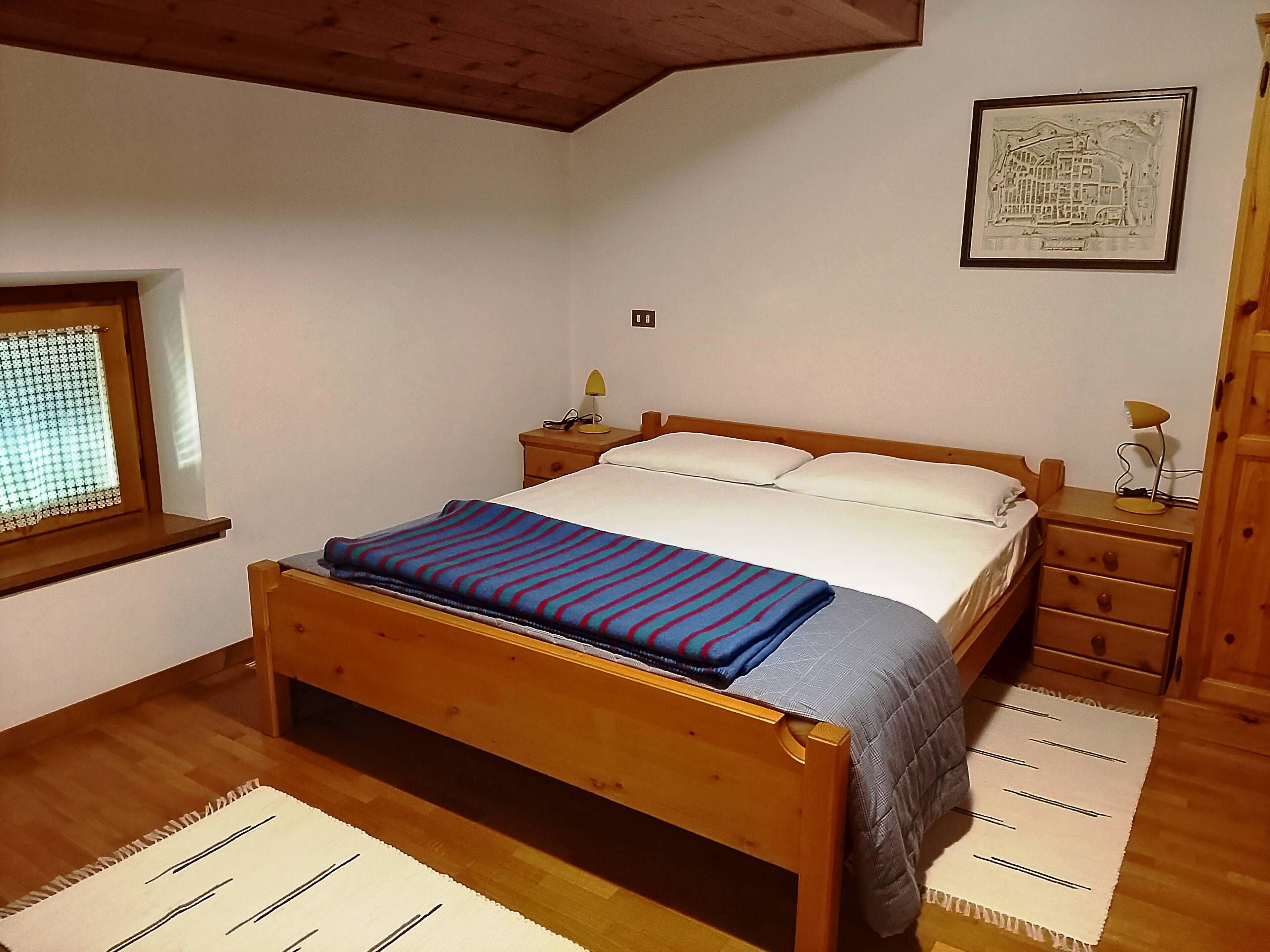 Un dormitorio con una cama de madera, una mesita de noche con lámpara, una ventana con vistas y un cuadro en la pared.