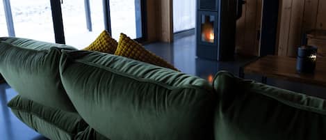A cozy room with a green sofa, a wood-burning stove, and a view of the snowy outdoors through large windows.