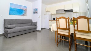 A compact living space with a grey sofa, wooden dining table with chairs, a kitchen area with white cabinets, and a painting of a coastal scene on the wall. - Beachfront apartment fully equipped with terrace and seaview, parking included. (Okrug Gornji)