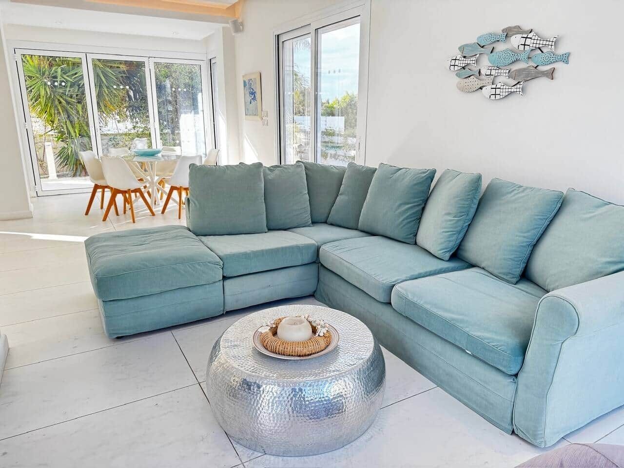 A modern living room with a light blue sectional sofa, a round glass coffee table, and a dining area with a white table and wooden chairs.