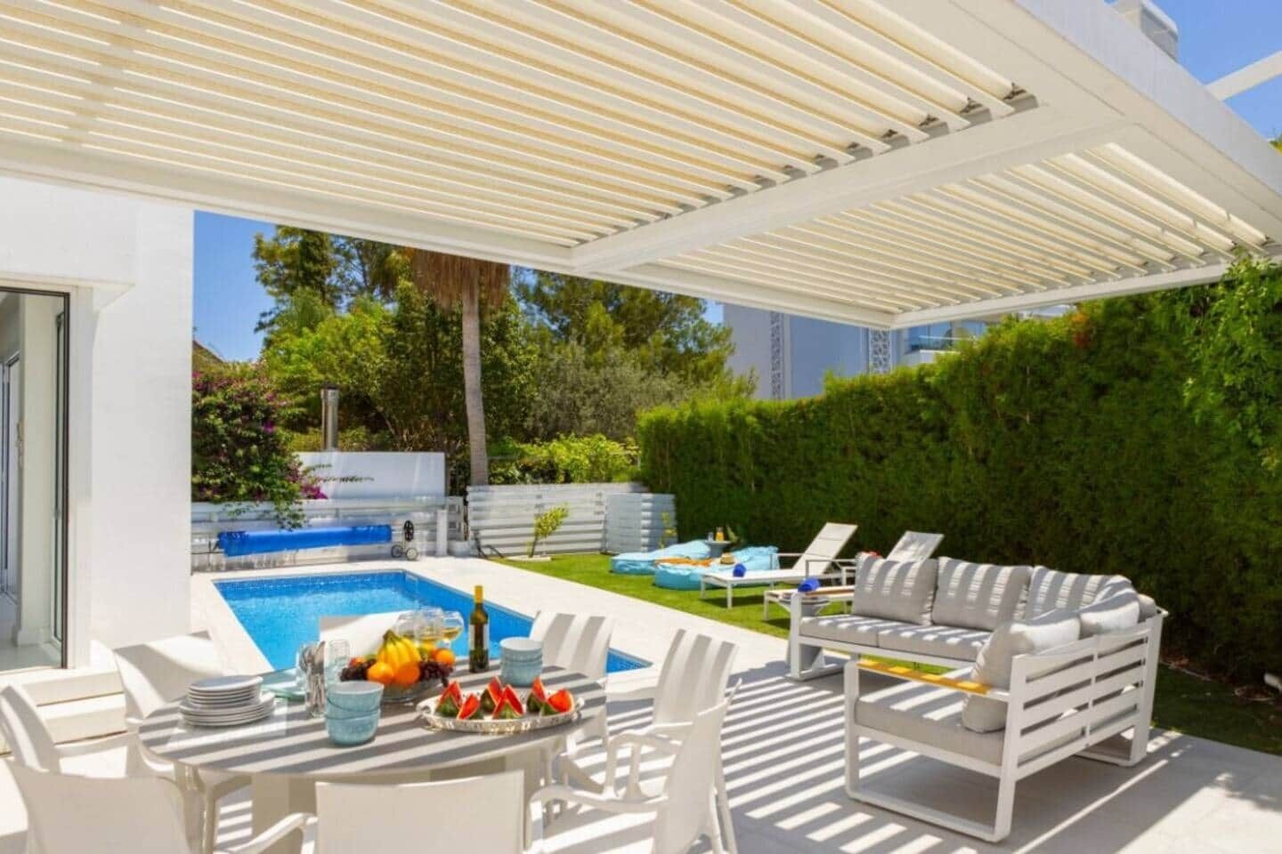 A modern outdoor pool area with a covered patio, lounge chairs, and a table set with fruits and drinks.