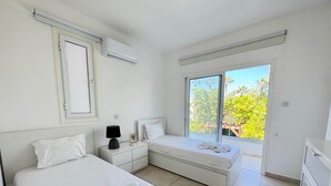 Quarto com duas camas, uma mesa de cabeceira, ar condicionado, janela com vista para árvores e uma porta deslizante.