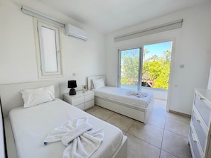 A bedroom with two beds, a nightstand, an air conditioner, a window with a view of trees, and a sliding door. - Protaras Seaside Serenity - Konnos Beach (Paralimni)