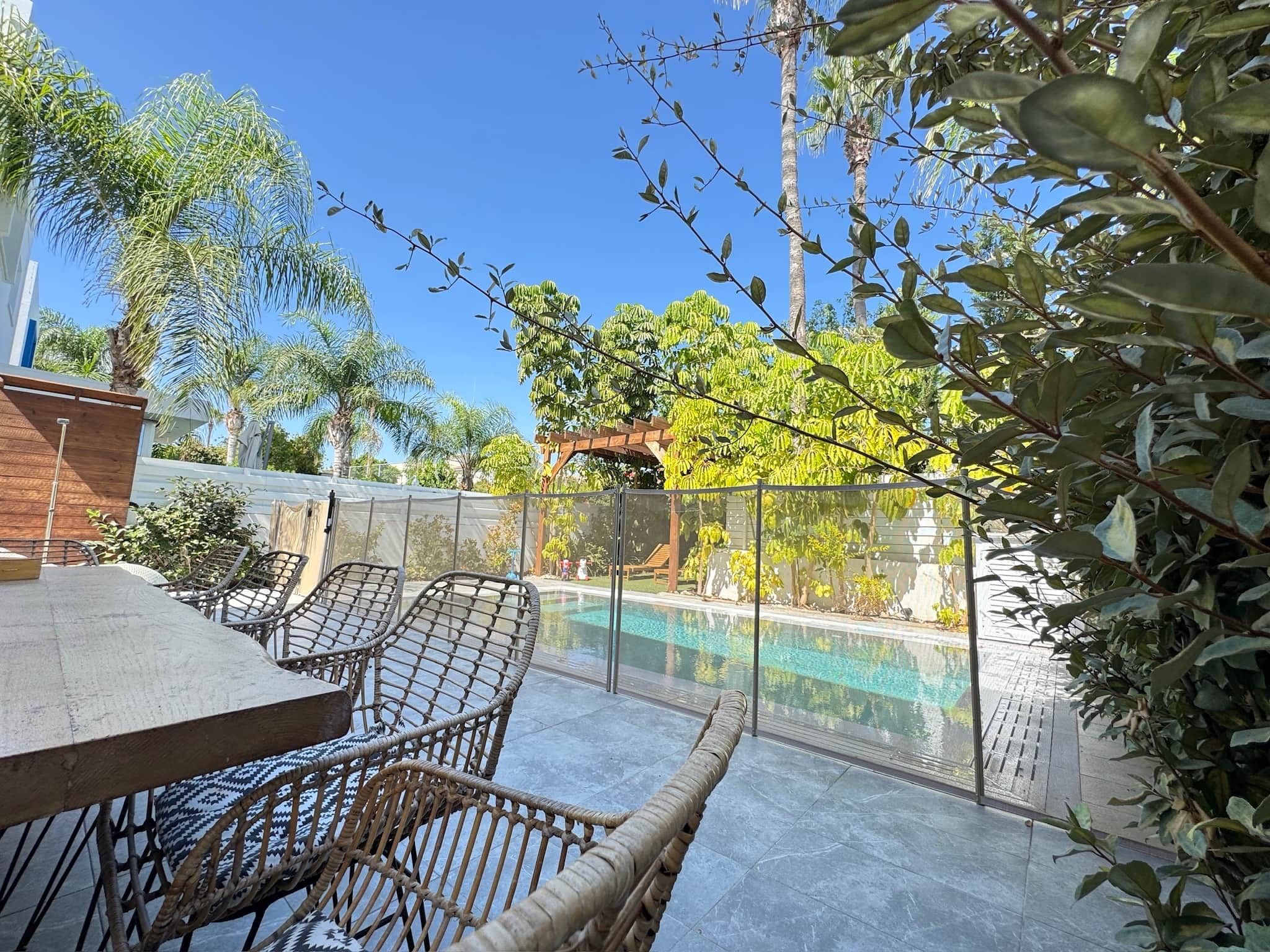 A pool area with a table and chairs, surrounded by greenery and a clear sky.