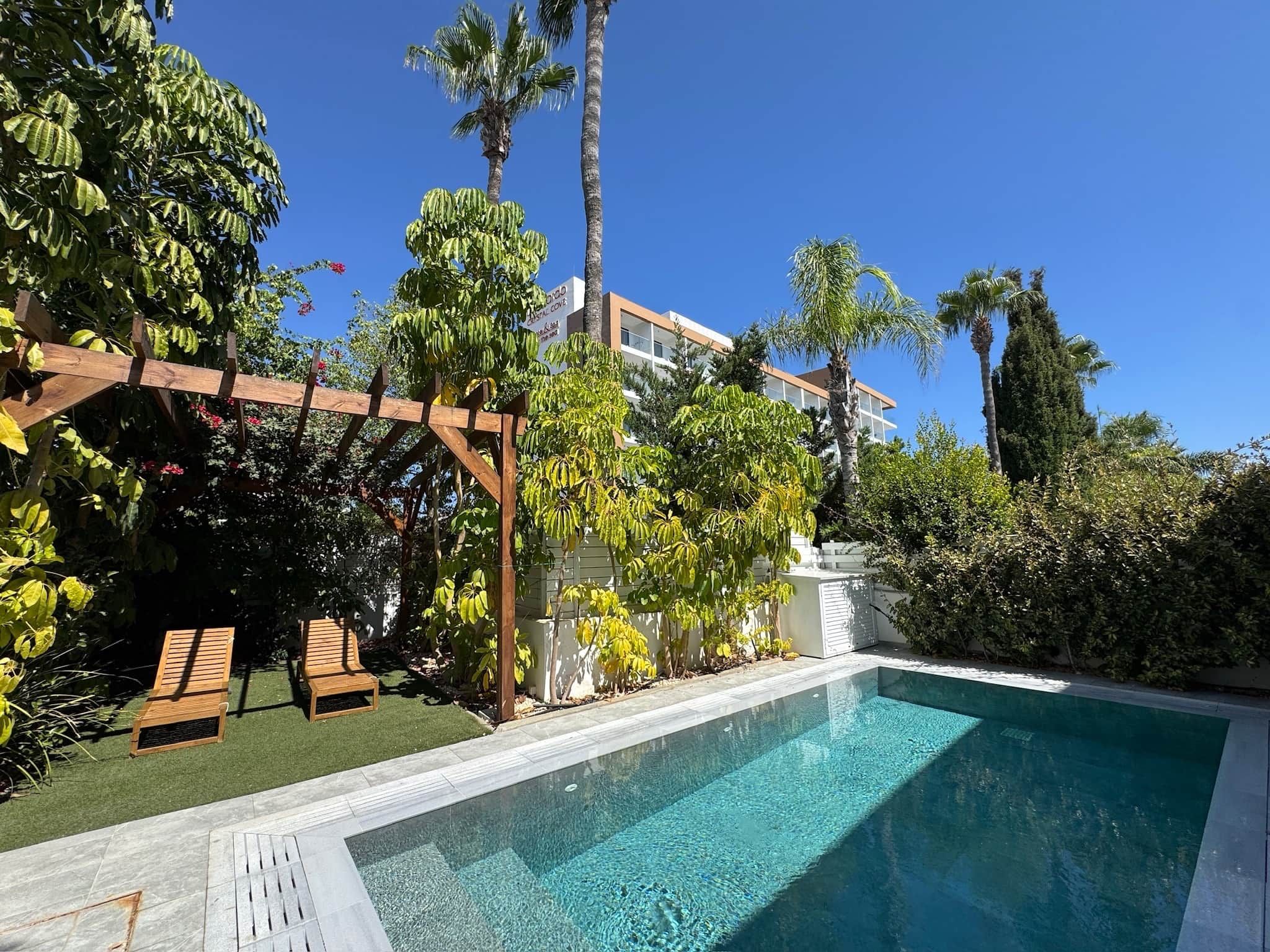 Une piscine agrémentée d’une pergola en bois et de chaises longues, entourée d’une végétation luxuriante et de grands palmiers.