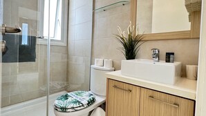 Uma casa de banho moderna com cabine de duche em vidro, sanita com assento estampado a verde e branco, lavatório com artigos de higiene e um armário de madeira.