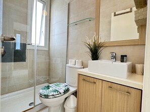 A modern bathroom with a glass shower enclosure, a toilet with a green and white patterned seat, a sink with toiletries, and a wooden cabinet. - Protaras Seaside Serenity - Konnos Beach (Paralimni)