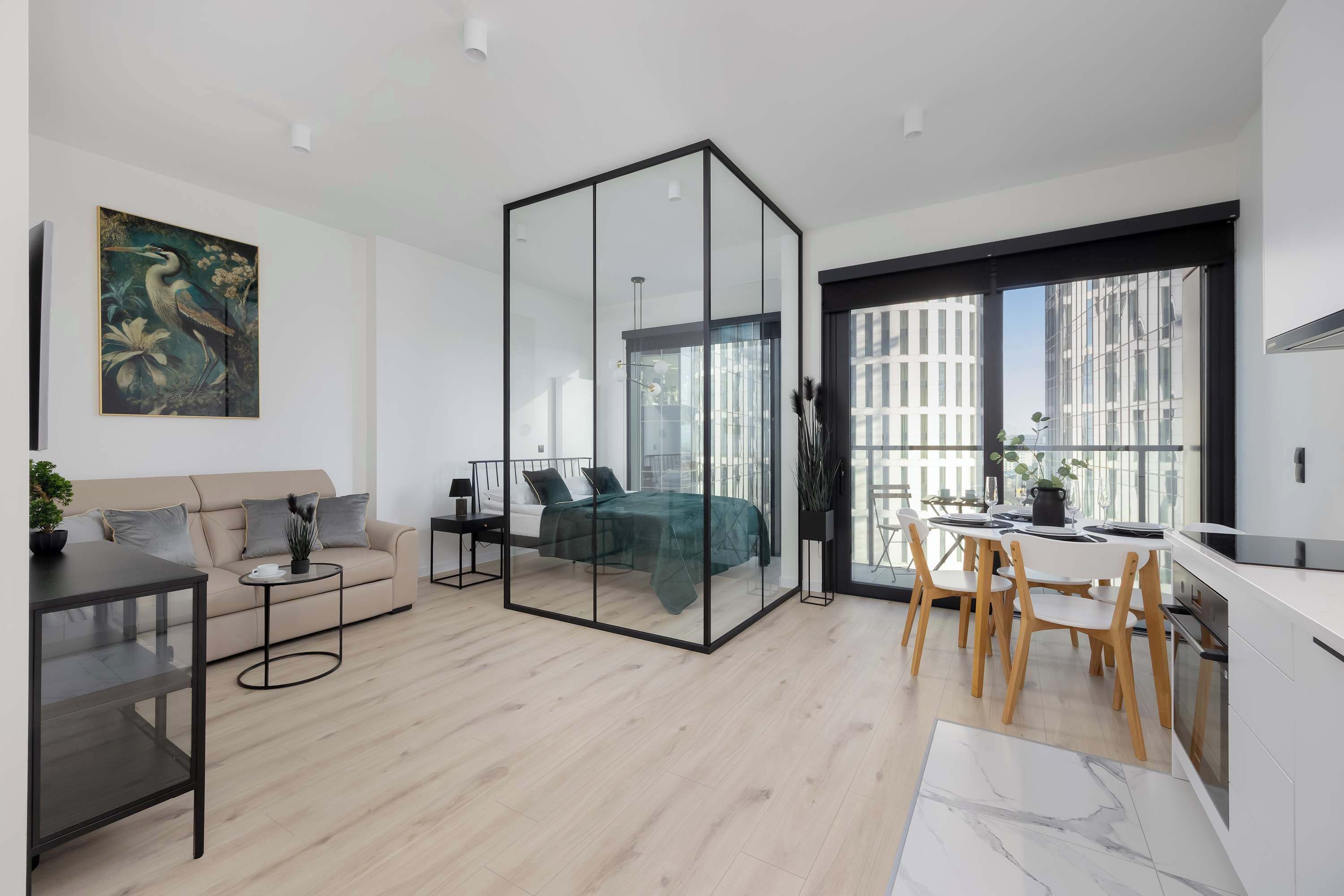A modern apartment with a glass partition separating the living area and bedroom. It includes a sofa, a dining table with chairs, a kitchen area, and a large window offering a city view.