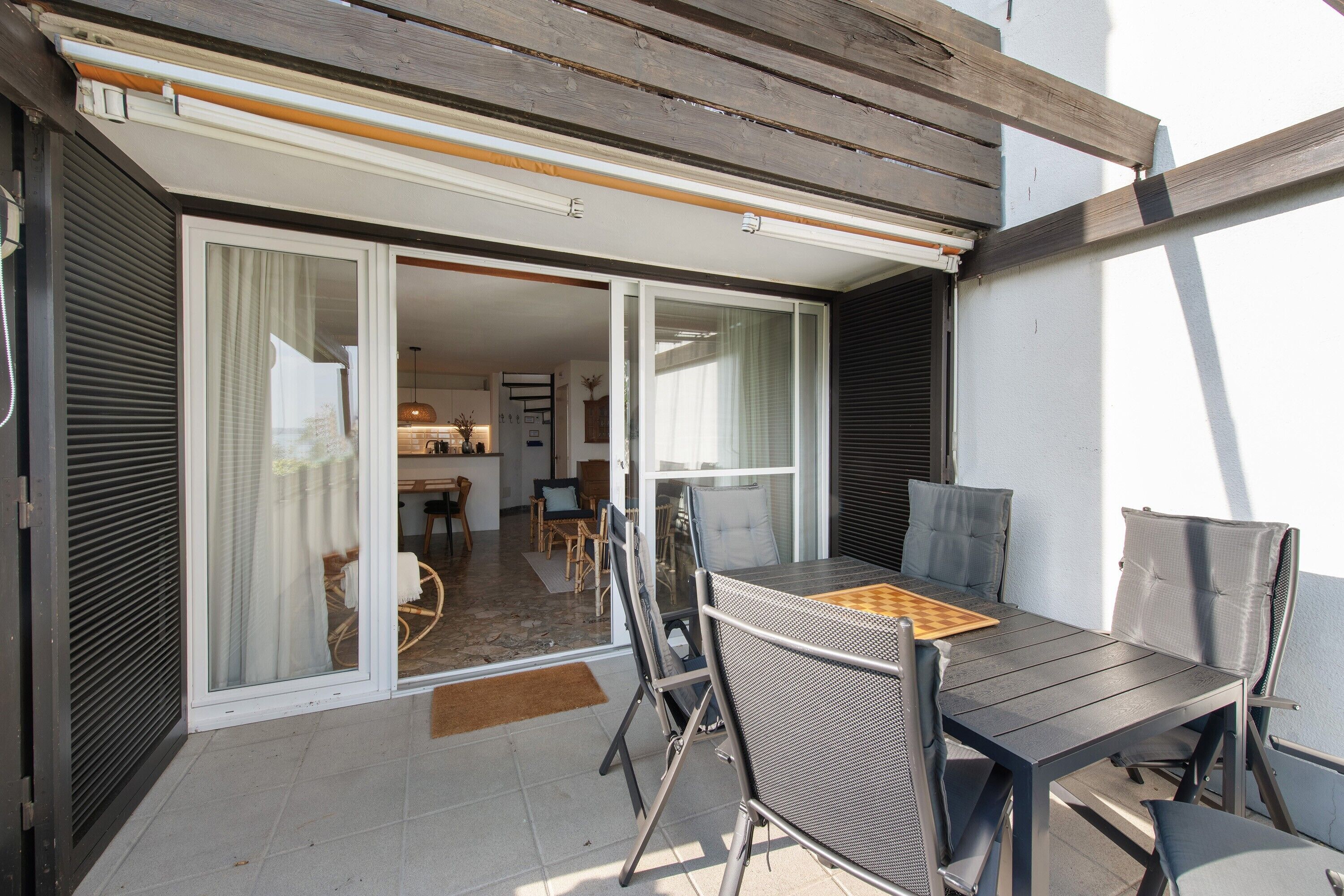 Eine Terrasse mit Tisch, Stühlen und Schachbrett, sowie eine Glasschiebetür zu einem Innenbereich mit Küche und Essbereich.