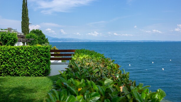 A lakeside view with a bench, greenery, and a distant mountain range. - Holiday Home 'Casa Liner - Steps From The Lake' with Lake View, Private Terrace and Wi-Fi (Moniga del Garda)