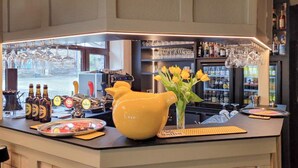 Un coin bar avec un grand pichet jaune, un vase rempli de fleurs jaunes, ainsi qu’une variété de bouteilles et de verres disposés sur le comptoir.