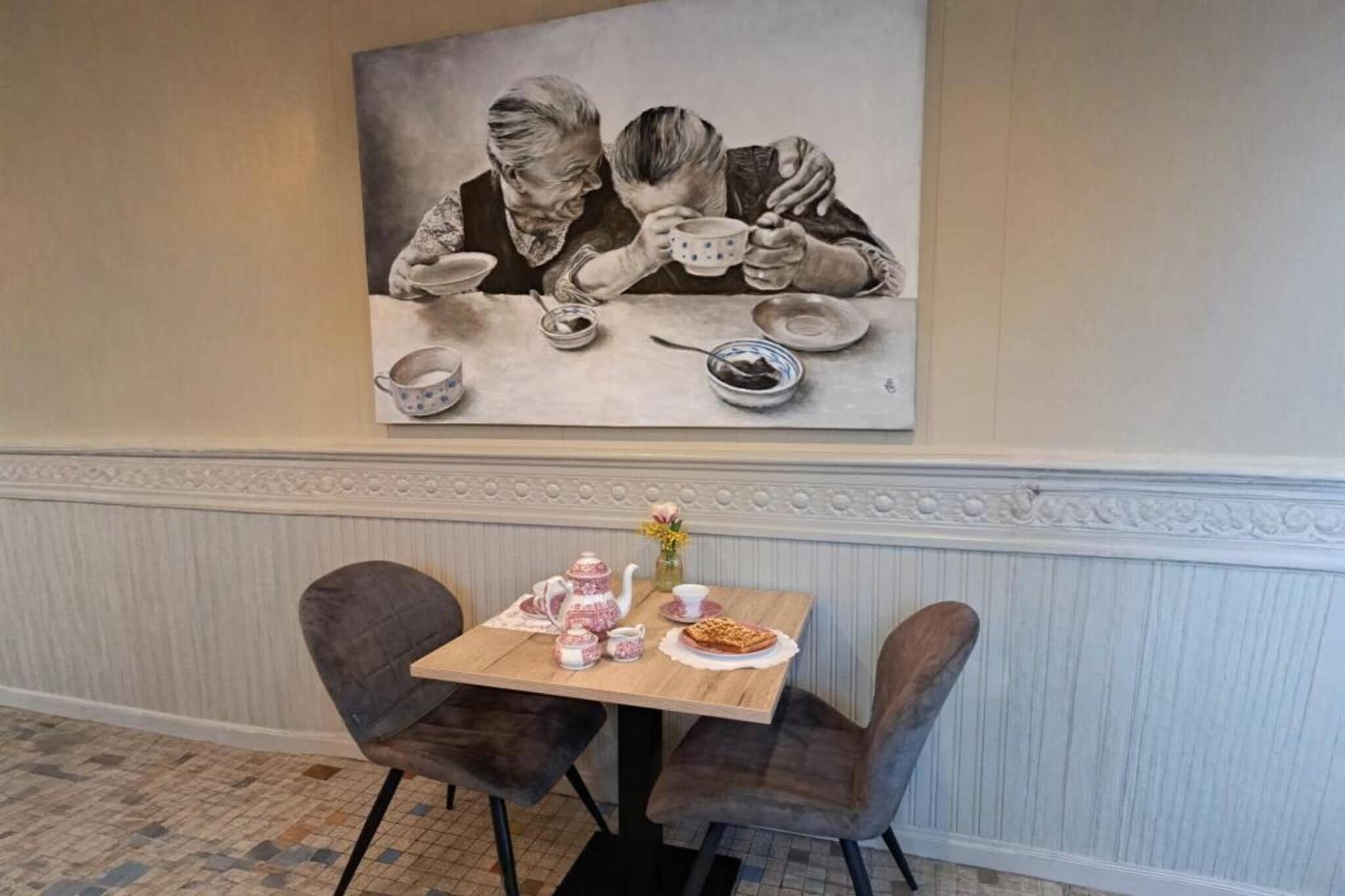 Een eethoek met een tafel gedekt voor twee personen, een schilderij van twee mensen die samen eten, en een vloer met een patroon.