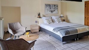 Une chambre à coucher comprenant un lit, deux chaises, une table de chevet, une lampe, une œuvre d’art murale et une porte.