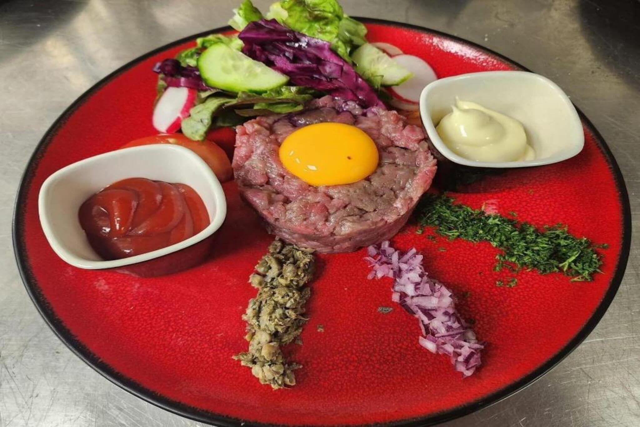 A plate with a raw meat dish topped with an egg yolk, accompanied by a side salad, ketchup, and a small bowl of sauce.