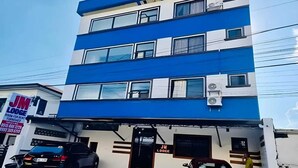 Un immeuble à plusieurs étages, doté d’une façade bleue et blanche, avec une enseigne indiquant « JM Lodge », ce qui signifie qu’il y a des chambres à louer, et deux voitures garées devant.