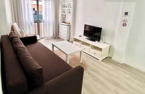 A living room with a brown sofa, a white coffee table, a TV on a white stand, and a white cabinet with shelves.