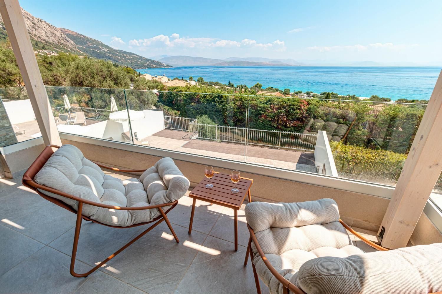 Un balcon agrémenté de deux fauteuils confortables et d’une petite table, offrant une vue imprenable sur un paysage côtier, avec la mer et les montagnes lointaines en toile de fond.