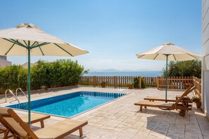 A rectangular pool with a wooden deck, two lounge chairs, and two umbrellas. - Villa Alya View in Mparmpati (Corfu)