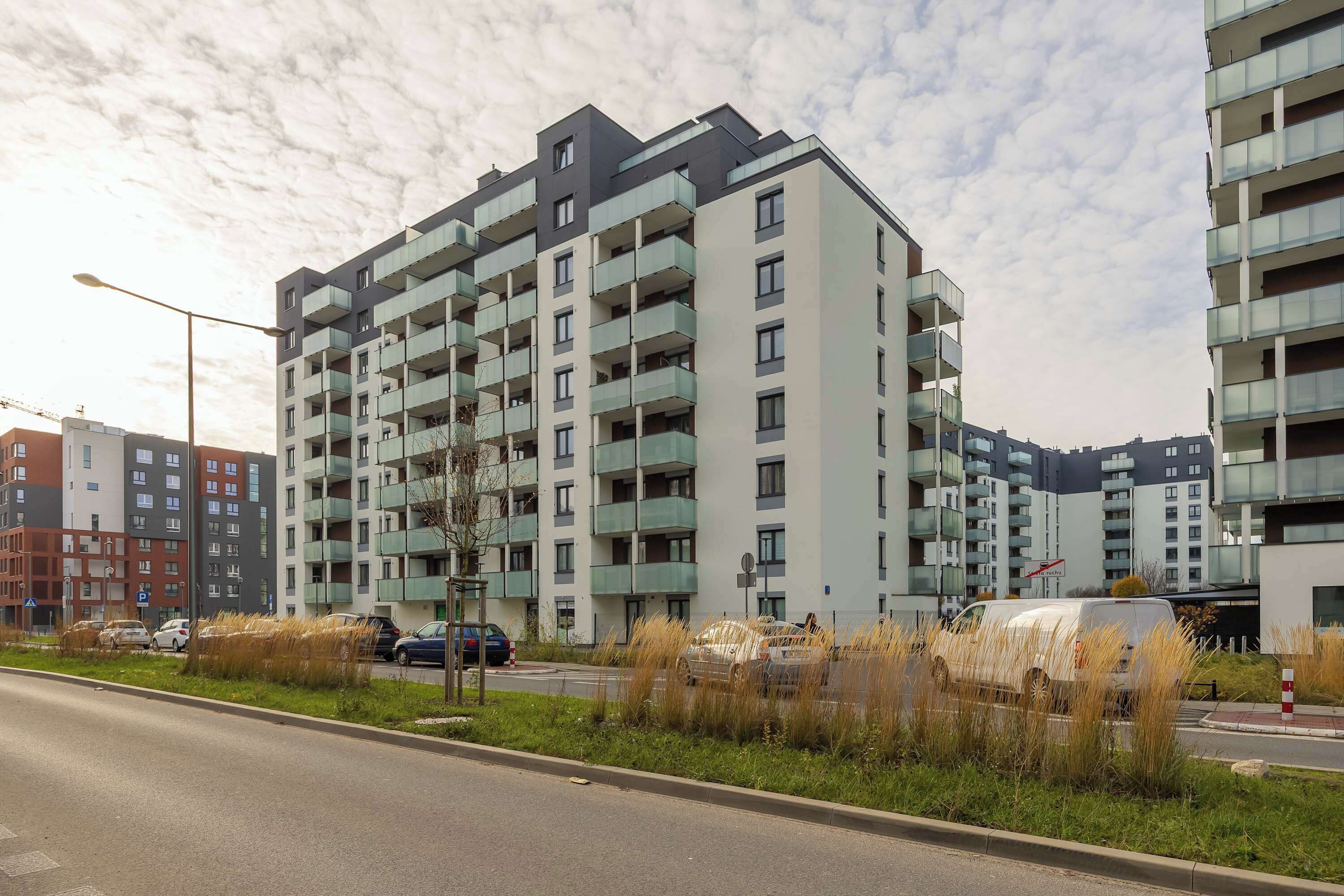 A multi-story residential building with balconies, a parking area, and a street with vehicles.
