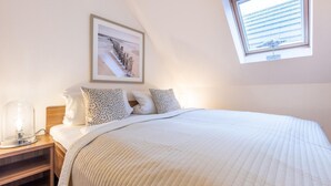 A bedroom with a large bed, a nightstand with a lamp, a framed picture on the wall, and a window with blinds. - Due/05a - Im Dünengrund App. 05a (Sylt)