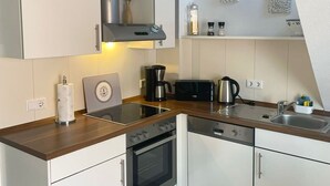 A modern kitchen with white cabinets, a stainless steel range hood, a built-in oven, a coffee maker, and a sink. - Due/05a - Im Dünengrund App. 05a (Sylt)
