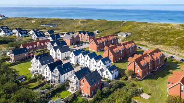 An aerial view of a residential area with houses, some with sloped roofs and others with flat roofs, surrounded by greenery and a clear view of the sea. - Due/05a - Im Dünengrund App. 05a (Sylt)