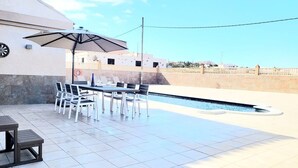 Un espace aménagé au bord de la piscine, avec une table et des chaises, un grand parasol et un sol carrelé.