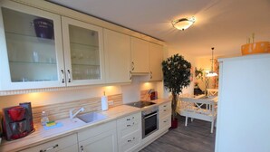 A modern kitchen with white cabinets, a sink, a stove, and a dining area with a white table and chairs. - DUE/09, Apartment 15 - Im Dünengrund App. 14 and 15 (Sylt)