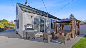 Ein kleines Haus mit Terrasse, Grill, Korbmöbeln und einer überdachten Sitzgelegenheit.
