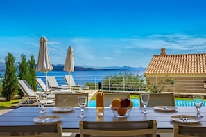 A dining table with chairs, a bottle of wine, and a bowl of fruit. There are two umbrellas and a pool in the background. - Villa Odysseus in Ionian Islands (Corfu)