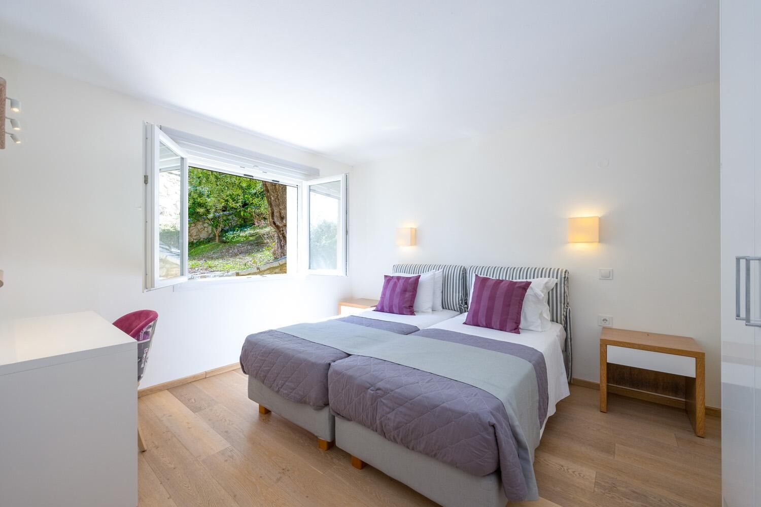 Ein Schlafzimmer mit einem großen Bett, zwei Nachttischen, einem Fenster mit Blick auf Grünflächen und einem kleinen Schreibtisch mit Stuhl.