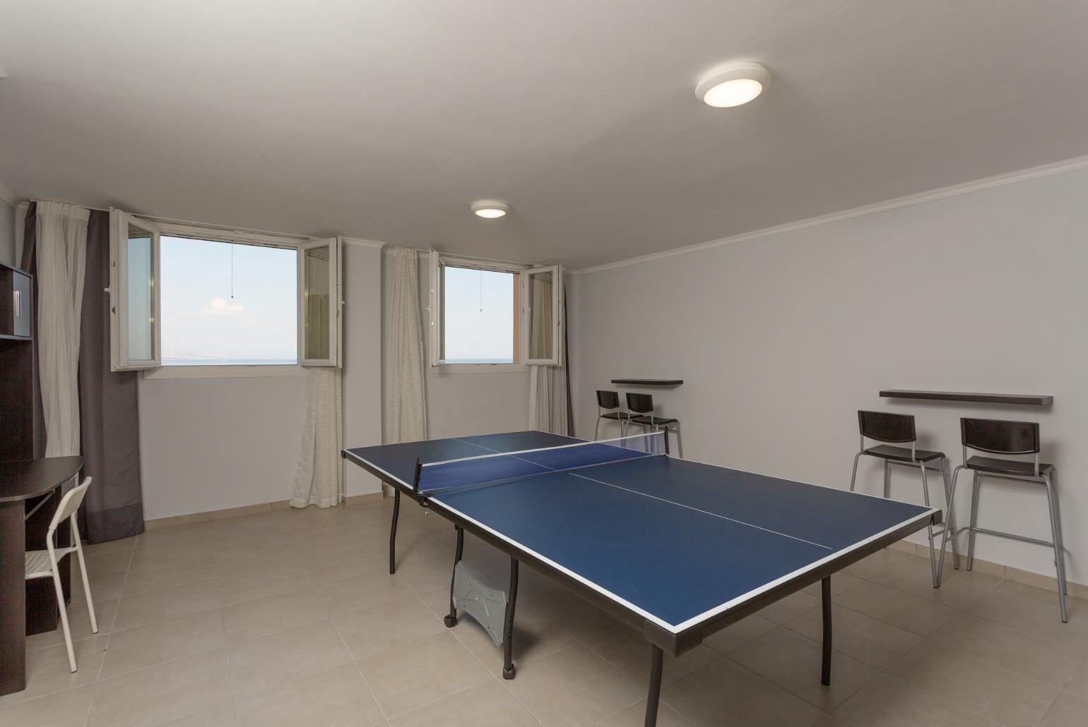 Une pièce avec une table de ping-pong, deux chaises, une petite table et une fenêtre avec des rideaux.