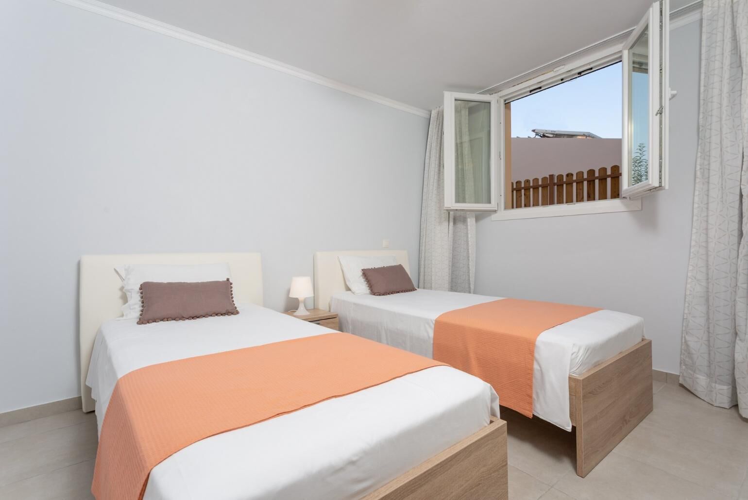 A hotel room with two beds, a window with a view of a fence, and a wooden floor.