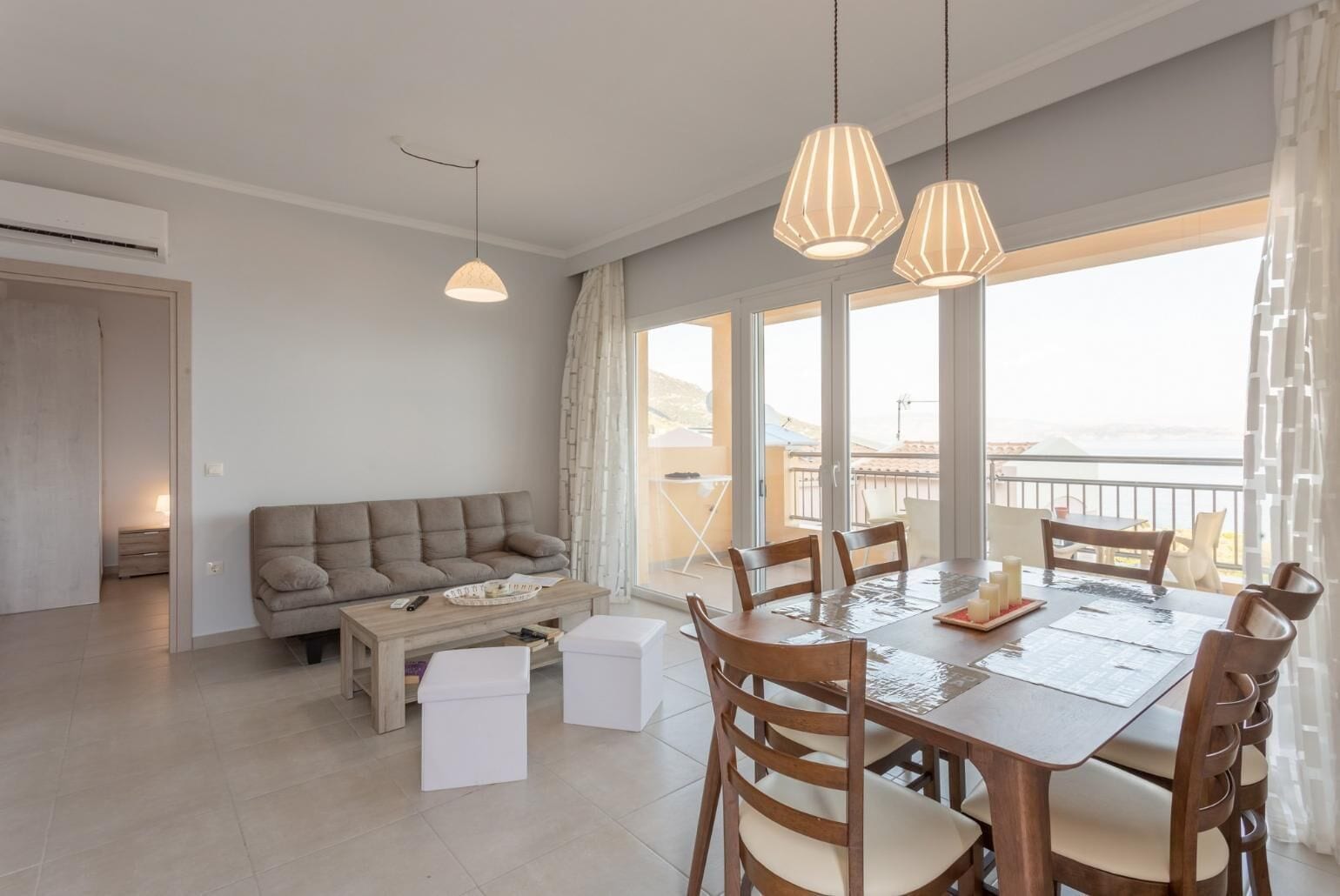 En moderne stue med sofa, sofabord og spisebord med stole. Store vinduer og en balkon med udsigt.