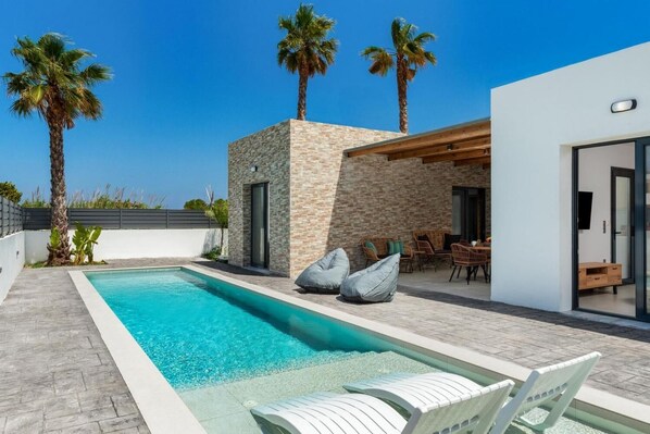 Un área de piscina moderna al aire libre con un muro de piedra, palmeras y mobiliario exterior.