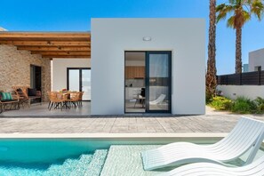 Una casa moderna con piscina, mobiliario exterior y una cocina visible a través de un ventanal.