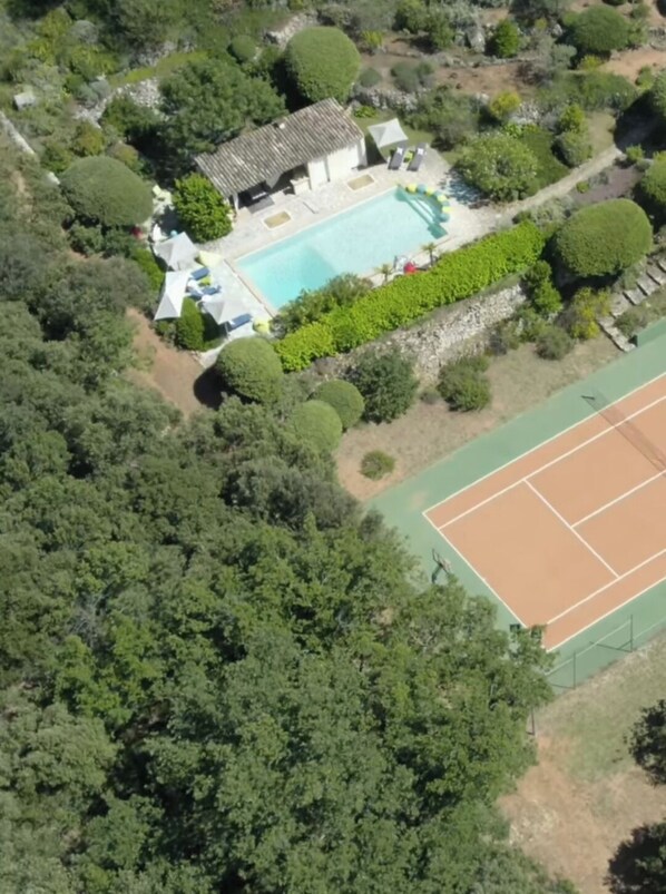 An aerial view of a property with a swimming pool, tennis court, and lush greenery.