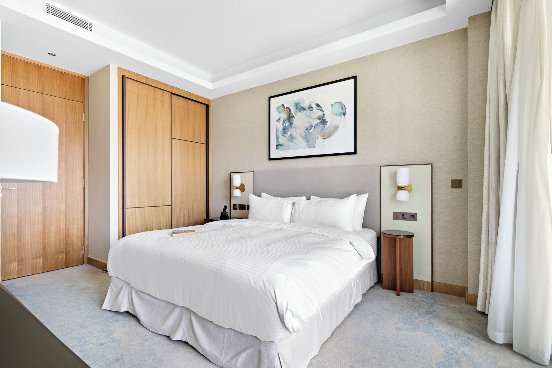 A modern hotel room with a large bed, wooden wardrobe, two bedside tables with lamps, and a framed abstract painting on the wall.