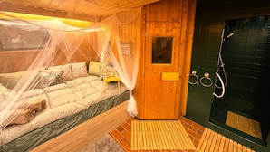 Ein Zimmer aus Holz mit einem Bett unter einem Moskitonetz, einer Holztür und einem Duschbereich mit Handtuchhalter.
