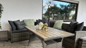 Ein Sitzbereich im Freien mit Korbmöbeln, einem Holztisch und Kissen, einer dekorativen Vase und Blick auf Bäume durch ein Fenster.
