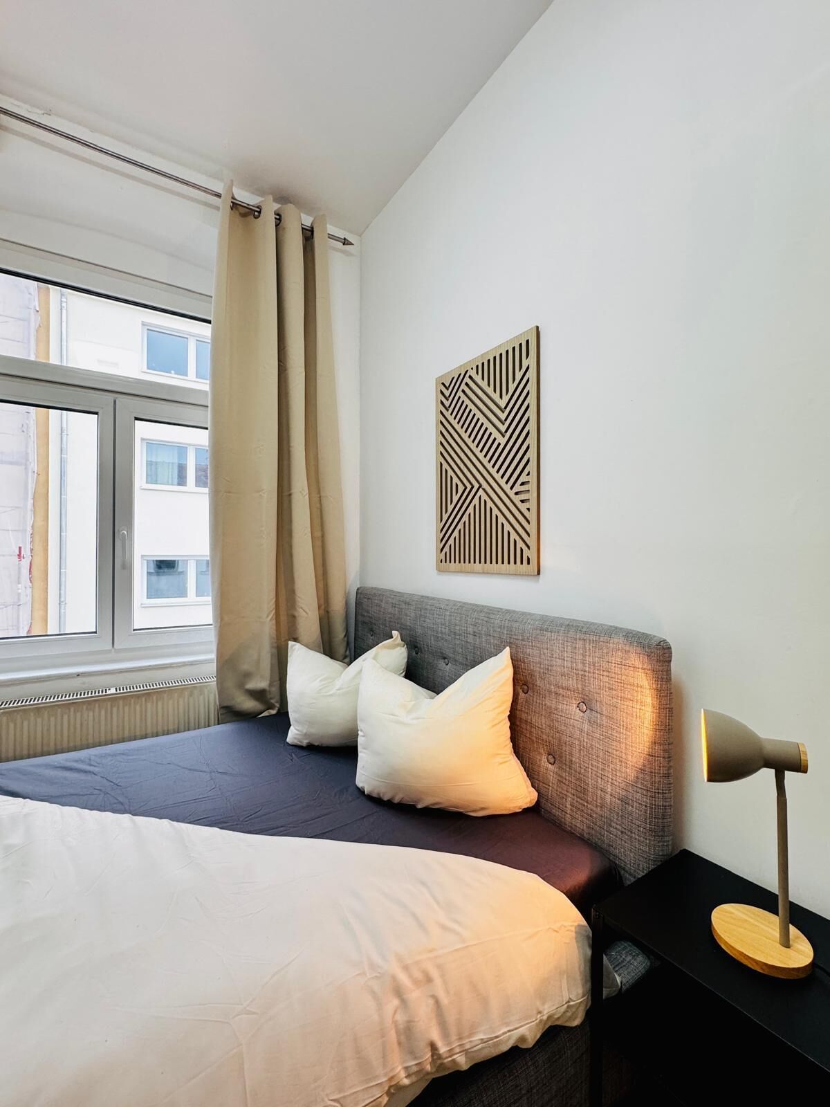 Ein Hotelzimmer mit Bett, Nachttisch, Lampe, einem Fenster mit Vorhängen und Wanddekoration.