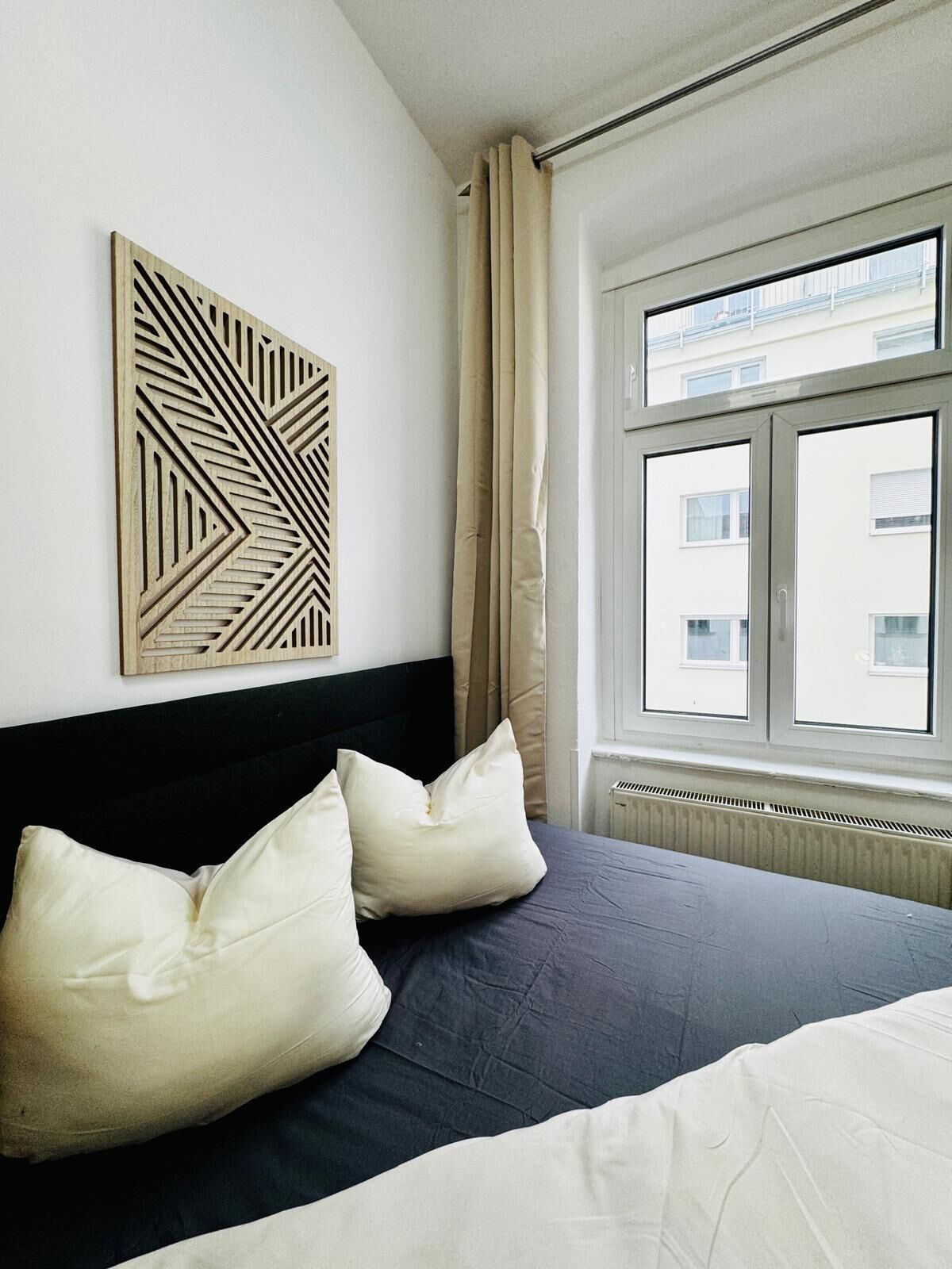A bedroom with a bed, two pillows, a geometric wall art piece, a window with curtains, and a radiator.