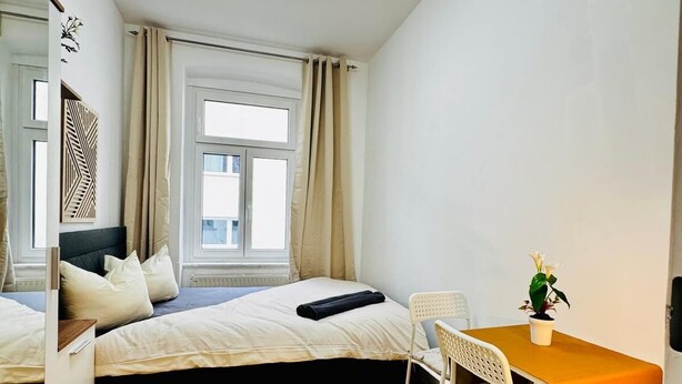 Ein gemütlicher Raum mit einem Bett, einem kleinen Tisch mit Stuhl, einer Zimmerpflanze und einem Fenster mit Vorhängen.